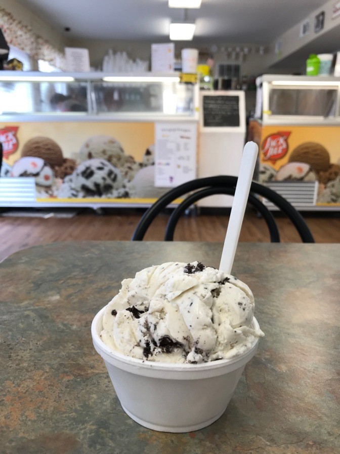 Cup of cookies and cream ice cream on table, with ice cream counter in background.