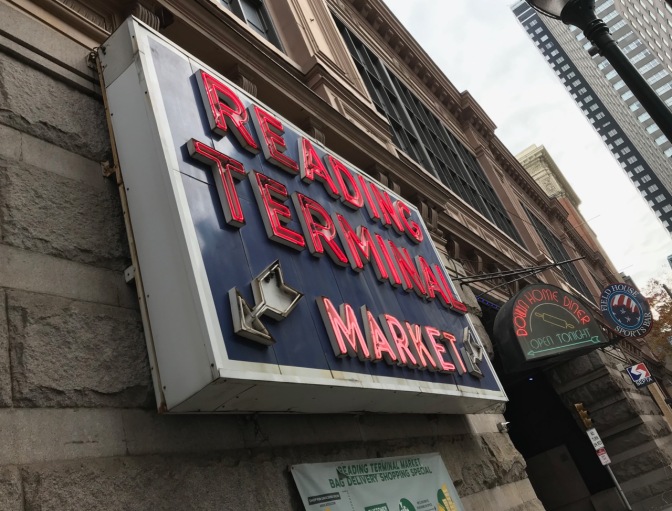 Exterior of Reading Terminal Market with neon sign that says READING TERMINAL MARKET on stone wall.