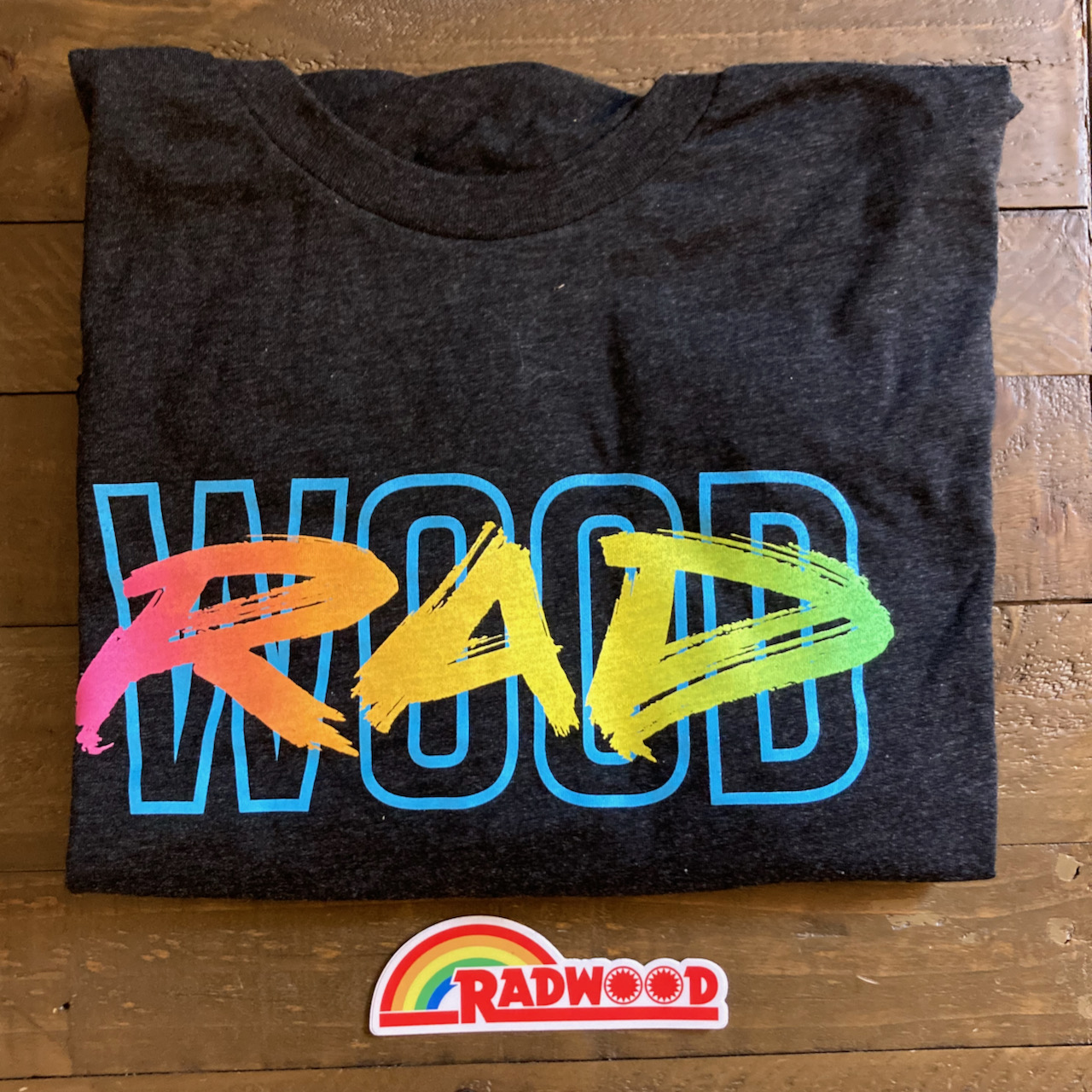 RADWOOD T-Shirt and Redwood rainbow sticker on wooden table..